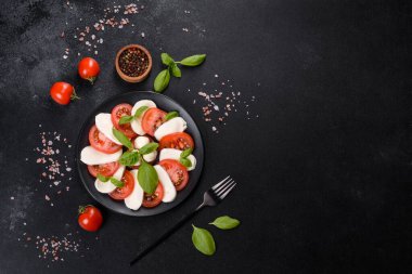 Dilimlenmiş domatesli İtalyan caprese salatası, mozzarella peyniri, fesleğen, zeytinyağı. Eskimiş siyah plaka ile koyu beton arka planda servis ediliyor.