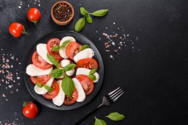 Dilimlenmiş domatesli İtalyan caprese salatası, mozzarella peyniri, fesleğen, zeytinyağı. Eskimiş siyah plaka ile koyu beton arka planda servis ediliyor.