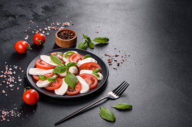Dilimlenmiş domatesli İtalyan caprese salatası, mozzarella peyniri, fesleğen, zeytinyağı. Eskimiş siyah plaka ile koyu beton arka planda servis ediliyor.