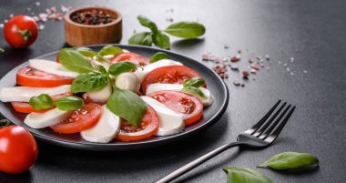 Dilimlenmiş domatesli İtalyan caprese salatası, mozzarella peyniri, fesleğen, zeytinyağı. Eskimiş siyah plaka ile koyu beton arka planda servis ediliyor.