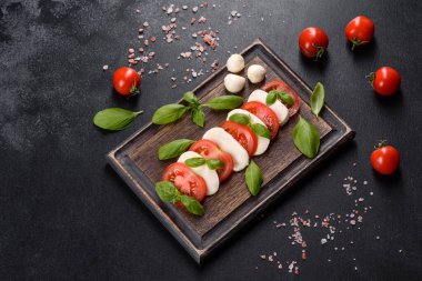 Dilimlenmiş domatesli İtalyan caprese salatası, mozzarella peyniri, fesleğen, zeytinyağı. Eskimiş siyah plaka ile koyu beton arka planda servis ediliyor.