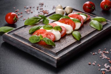Dilimlenmiş domatesli İtalyan caprese salatası, mozzarella peyniri, fesleğen, zeytinyağı. Eskimiş siyah plaka ile koyu beton arka planda servis ediliyor.