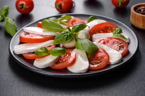 Dilimlenmiş domatesli İtalyan caprese salatası, mozzarella peyniri, fesleğen, zeytinyağı. Eskimiş siyah plaka ile koyu beton arka planda servis ediliyor.