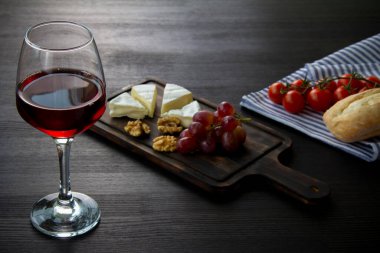 Bir bardak kırmızı şarap, peynir, üzüm, tahta kesme tahtasında ceviz. Peçetede ekmek rulosu ve kiraz domatesleri. Karanlık arka plan. Romantik akşam yemeği konsepti. Üst görünüm