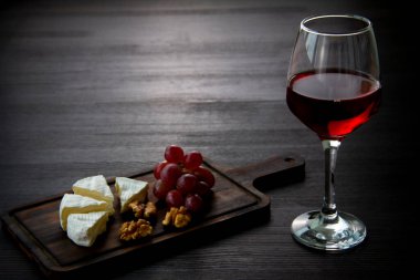 Bir bardak kırmızı şarap, peynir, üzüm, tahta kesme tahtasında ceviz. Karanlık arka plan. Romantik akşam yemeği konsepti. Üst Manzara. Boşluğu kopyala
