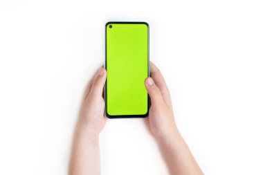 Küçük bir çocuk, beyaz arka planda izole edilmiş, boş yeşil ekranlı bir akıllı telefonu iki elle tutuyor. Üst görünüm, alanı kopyala