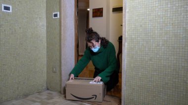 Avrupa, İtalya, Milan Aralık 2020 - Maskeli kadın, Covid-19 Coronavirus 'un kilit altına alındığı sırada evinin kapısındaki dairede Noel için Amazon paketi aldı