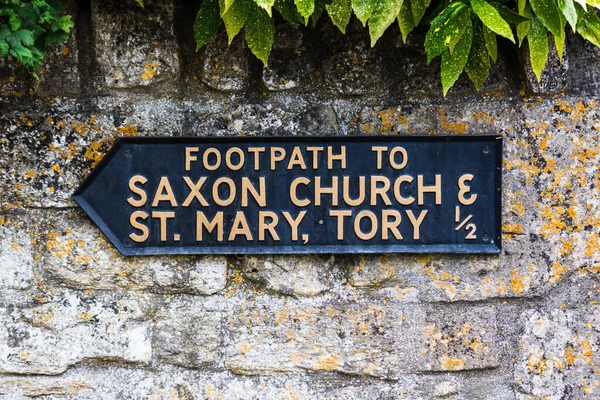 A black metal sign on a wall informs walkers of the footpath to the Saxon Church and St Mary, Tory in the historic town of Bradford on Avon