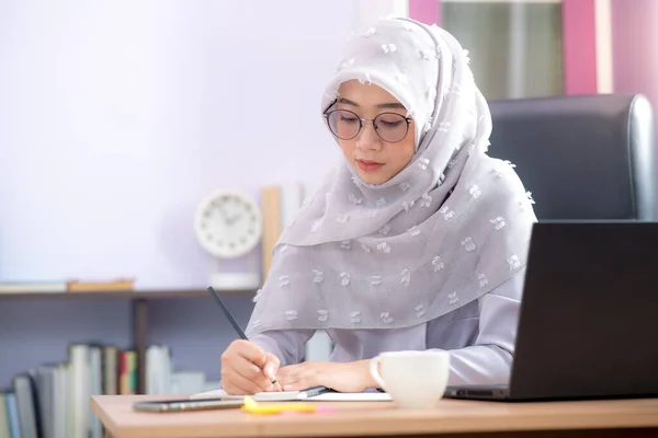 Asian Muslim woman office worker sitting in front of laptop computer at desk and smiling happy at office.