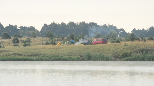 Fumo dal fuoco nel campo con tende e auto sulla riva del fiume — Video Stock