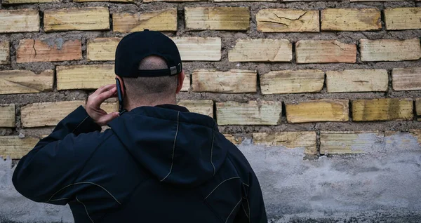man in a cap talks on the phone,with his back to a brick wall