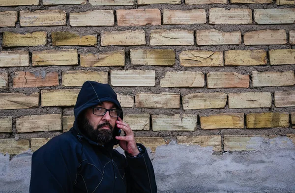 hooded man talks on the phone, in front of a brick wall