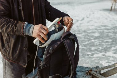 Biri buz tutmuş bir gölün arka planına karşı patenleri sırt çantasına koyar.