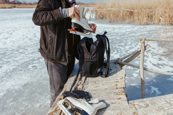 Biri buz tutmuş bir gölün arka planına karşı patenleri sırt çantasına koyar.