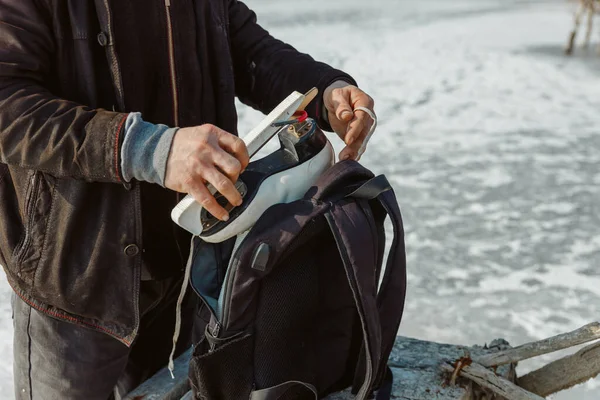 Biri buz tutmuş bir gölün arka planına karşı patenleri sırt çantasına koyar.