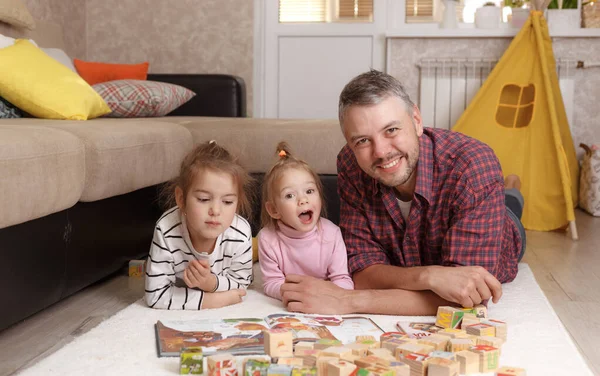 happy father in a plaid shirt playing with his daughters with wooden blocks lying on the floor. Family games with children during the pandemic.