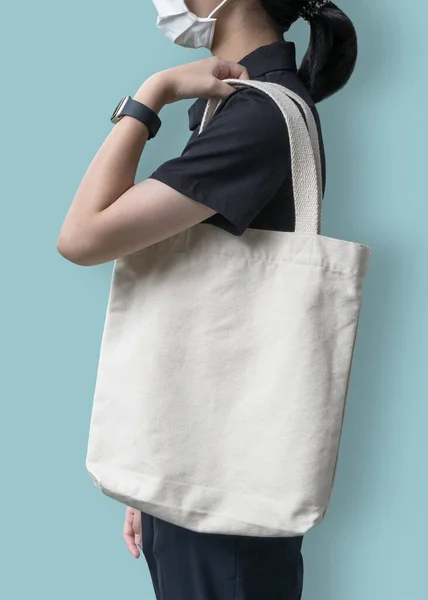 Tote bag mockup with canvas white cotton fabric cloth for eco shopping handbag mock up blank template on shoulder of girl with face mask isolated on pastel blue background (clipping path)