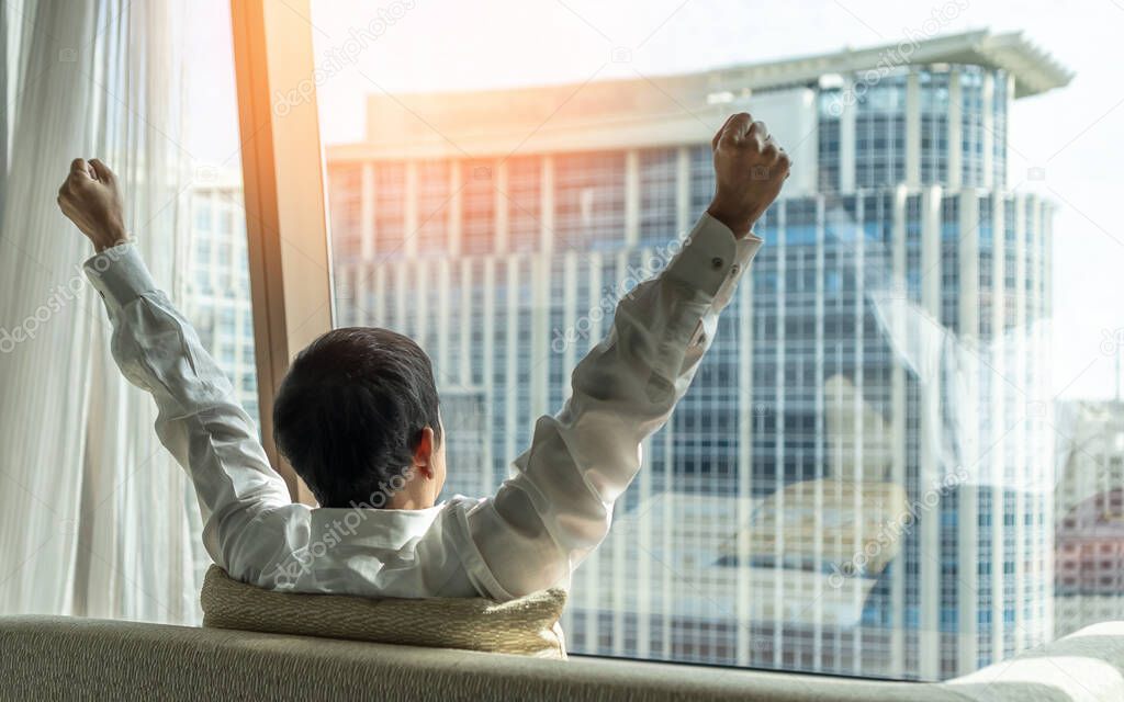 Business achievement ambition concept with businessman raising fists happily, relaxing in office or hotel room, resting and looking forward to city building urban scene 