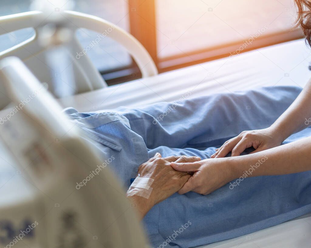 Caregiver and elderly senior patient (aged old adult person) holding hands in hospital bed or nursing hospice, geriatrician palliative home, while caretaker having  medical health care service