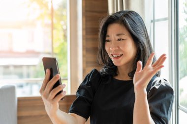 Kadın kullanıcı dalgaları sosyal medya uygulaması kullanarak cep telefonuyla selamlaşırken covid-19 sırasında sosyal mesafeler için yeni normal yaşam tarzında evde görüntülü yakınlaştırma toplantısı