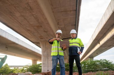 İnşaat uzmanları, şehir ortamında takım çalışması ve planlamayı vurgulayan bir köprü altında proje detayları üzerinde strateji oluşturuyorlar..