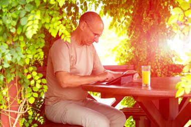 Casual dressed man with gadget and glass of orange juice sitting in wood arbor