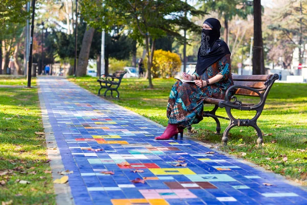 Young ισλαμική γυναίκα κάθεται σε παγκάκι στο πάρκο κατά μήκος πολύχρωμο λιθόστρωτο δρομάκι — Φωτογραφία Αρχείου