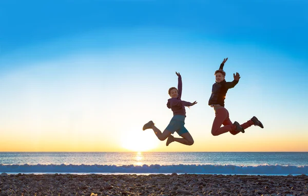 Holka a chlap skákat vysoké s rukama nahoru efektní východ slunce na pobřeží oceánu — Stock fotografie