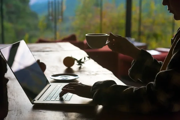 Silhouette einer am Computer arbeitenden Person, die Kaffee am Holztisch trinkt — Stockfoto