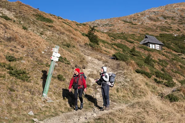 Dos excursionistas caminando en la ladera cubierta de hierba de montaña mirando Trail Sign —  Fotos de Stock