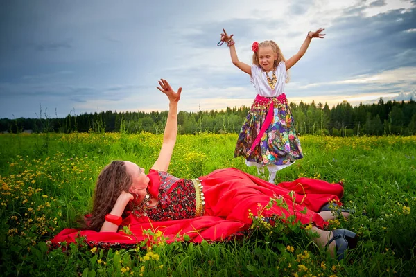 Mother and daughter in colorful Gypsy dresses resting and having fun in meadow with green grass. Little girl who is dancer or model dancing for adult woman looking as gypsy at summer evening on nature