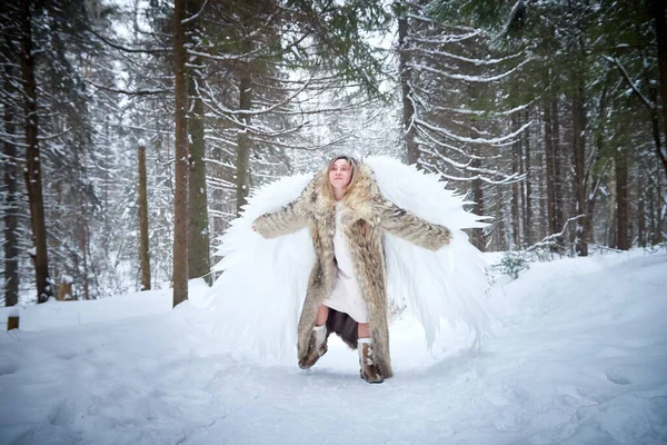 A middle-aged woman in a large warm fur coat and white angel wings in a winter forest with snow and snowdrifts. Fairy angel in cold forest in Russia before Christmas