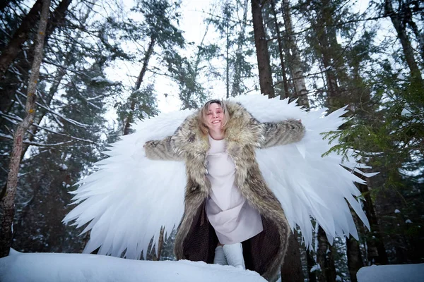 A middle-aged woman in a large warm fur coat and white angel wings in a winter forest with snow and snowdrifts. Fairy angel in cold forest in Russia before Christmas