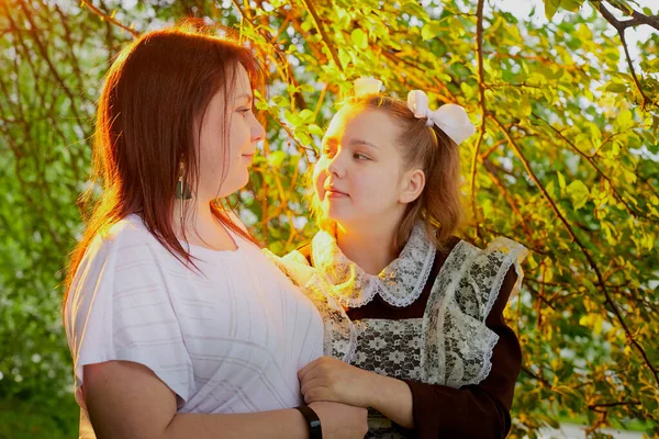 Portrait of young girl in an old school uniform of the USSR with black dress and a white apron with her mother in the park. Teenager and mom among the greenery after graduating from school in Russia