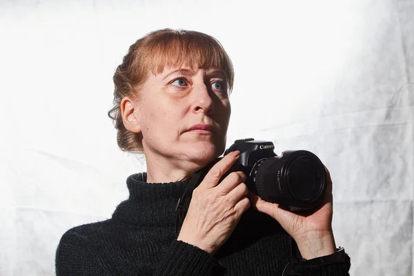 Female photographer in a studio in black dress with camera on white background. Woman posing indoors with cam