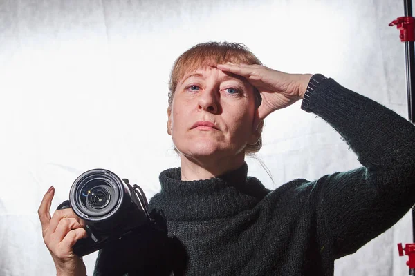 Female photographer in a studio in black dress with camera on white background. Woman posing indoors with cam