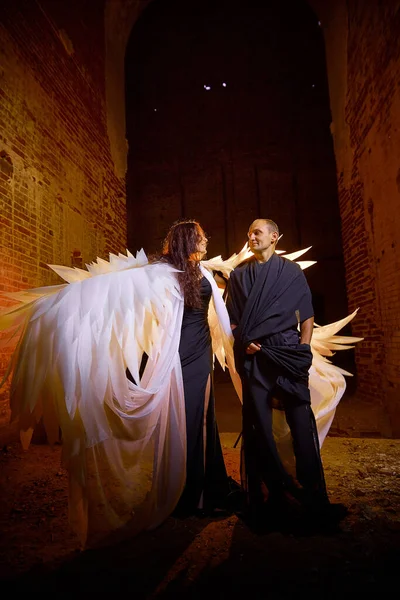 Couple in old dark clothes in abandoned castle with old brick walls. Man in black dress and woman with white wings posing in dark gloomy mysterious dangerous hall. The evil king and Queen on Halloween