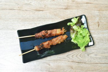  Baharatlı soslu ızgara domuz sarması, ahşap sopaya saplanmış ve tabağında sebze var.