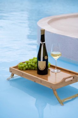 Bir şişe Beyaz Şarap ve Üzümler Lüks Yüzme Havuzu 'nda Yüzen Ahşap Tepside, Yaz Tatili Konsepti.