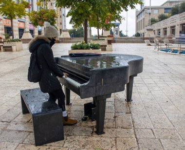 Kudüs, İsrail - 5 Kasım 2020: Koruyucu maske takan genç bir kadın, yağmurlu bir günde Kudüs, İsrail, belediye binası yakınlarında sokak piyanosu çalıyor.
