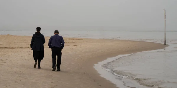 An elderly couple take a walk on a beach on a hazy morning.