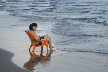 Tel Aviv, İsrail - 20 Ağustos 2021: Tel Aviv, İsrail 'de turuncu plaj sandalyesinde oturan bir kadın, şafak vakti akıllı telefonunu kullanıyor..