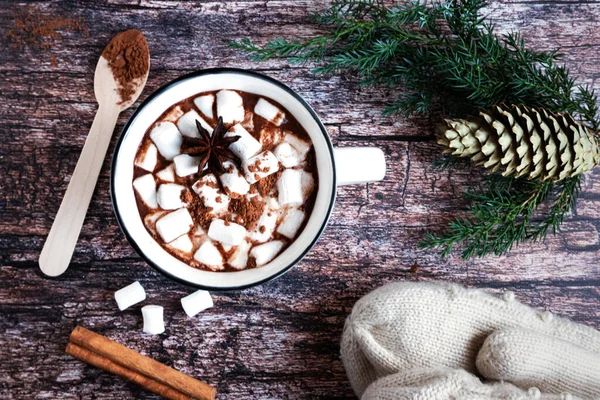 Vista superior de uma xícara de cacau quente ou chocolate com marshmallows em uma mesa de madeira rústica com ramos de abeto, cone de abeto, colher de madeira, canela e luvas de malha. — Fotografia de Stock