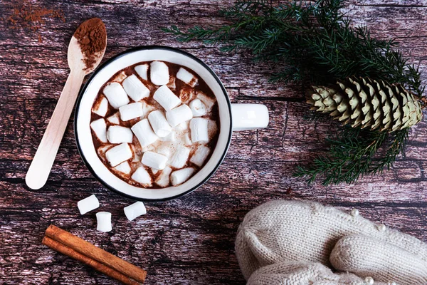 Vista superior de uma xícara de cacau quente ou chocolate com marshmallows em uma mesa de madeira rústica com ramos de abeto, cone de abeto, colher de madeira, canela e luvas de malha. — Fotografia de Stock