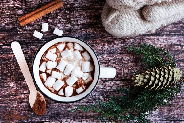 Vista superior de uma xícara de cacau quente ou chocolate com marshmallows em uma mesa de madeira rústica com ramos de abeto, cone de abeto, colher de madeira, canela e luvas de malha. — Fotografia de Stock