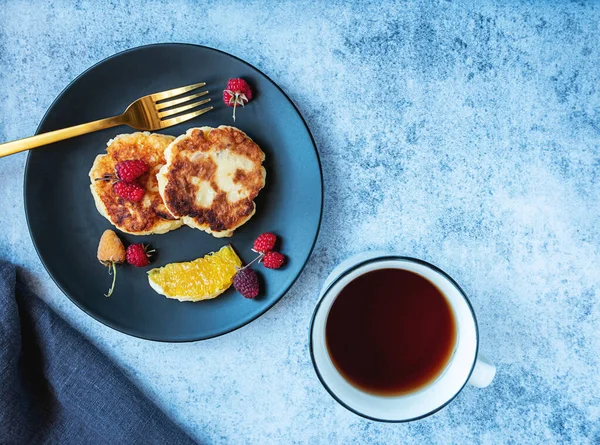 Panqueques de requesón, sirniki ruso, en un plato negro con bayas, naranja y taza de té. Desayuno saludable rico en calcio. Con espacio de copia. — Foto de Stock