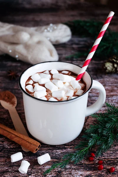 Weiße Tasse heißer Kakao oder Schokolade mit Marshmallows und einem gestreiften Papierstroh auf einer rustikalen Holztischplatte mit Tannenzweigen und gestrickten Handschuhen im Hintergrund. — Stockfoto