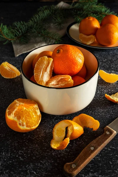 Tangerinas suculentas doces em uma tigela branca, parcialmente descascadas, com uma faca e um ramo de abeto no fundo escuro — Fotografia de Stock