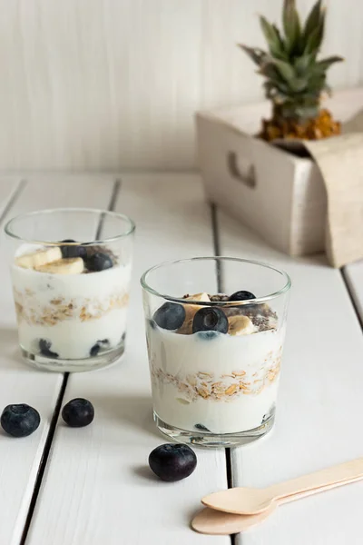 白い木製のテーブルの上に、層状ヨーグルト、オート麦、果物と2つのメガネ。健康的な朝食、健康的なライフスタイルのコンセプト — ストック写真