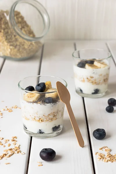 Dvě sklenice s vrstveným jogurtem, ovsem a ovocem, na bílém dřevěném stole. Zdravá snídaně, zdravý životní styl koncept — Stock fotografie